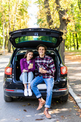 travel, summer vacation, road trip, leisure and people concept. Happy couple drinking coffee from disposable cups sitting on trunk of hatchback car outdoors