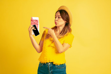 Happy holding tickets for traveling. Caucasian woman's portrait on yellow studio background. Beautiful model in cap. Concept of human emotions, facial expression, sales, ad. Summertime, travel, resort