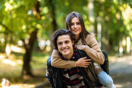 Man with stubble and headphones on neck carrying his beautiful woman in cap on back having journey trip, trendy stylish joyful couple hugging having meeting outdoors, sunny day