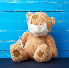 beige big teddy bear with patches sitting in a white medical mask in the paw of a syringe