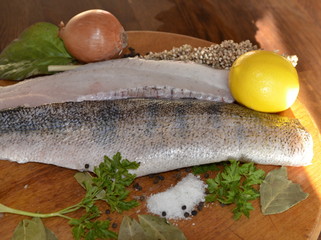 On a wooden board lies fresh pike perch fish fillet, yellow lemon, onion, salt, herbs, spices.