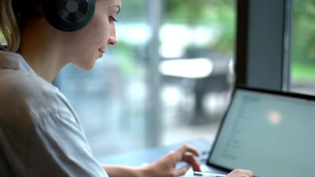 Caucasian girl in bluetooth headphones for noise cancelation typing message on laptop computer, young woman listening audio book via earphones during working on netbook