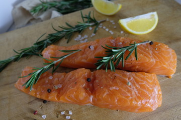 Two salmon or trout steaks lie on a wooden cutting board with fresh rosemary, chopped lemon, peppercorns and sea salt crystals. Horizontal orientation. Close-up.
