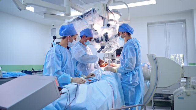 Group of surgeons performing operation to a patient. Doctors in medical uniform look into modern microscope during surgery in the operating room.