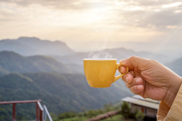 Hand holding a white cup of hot espresso coffee mugs and nature view of the mountain landscape in the morning with sunlight