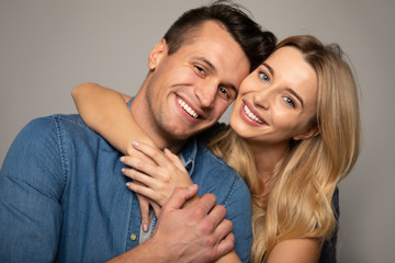 Radiant smiles. Close up photo of a blonde woman, who is hugging from the back her husband in a denim jacket, both are smiling and looking in the camera.
