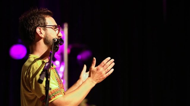 A slim man wearing glasses and tribal print t shirt is seen standing by a microphone stand. during a spoken word festival dedicated to native culture