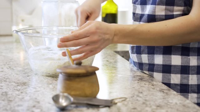 Woman preparing dough for baking bread while adding ingredients to dough and stirring in glass bowl