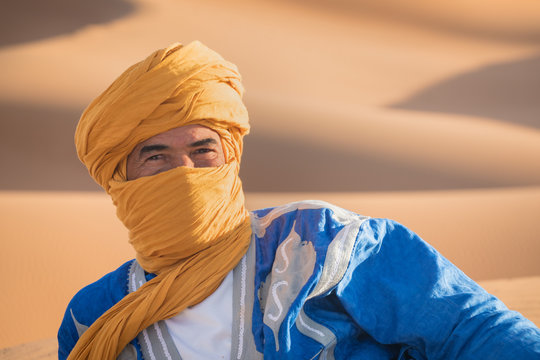 Bedouin nomad, Sahara desert, Morocco. Portrait of a Bedouin nomad with colorful turban and big smile sitting on sand dune popular tourist spot. A tuareg man portrait with his traditional clothes.