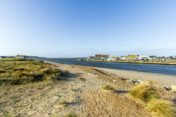 A view of a water channel between Christchurch (UK) harbor and bay with sandy beach and fishering buildings along the banks under a majestic blue sky