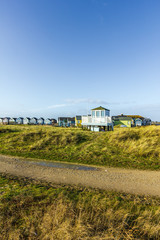 A view of colorful beach huts with a lifeguard post, track and grass in the foreground under a majestic blue sky and some white clouds