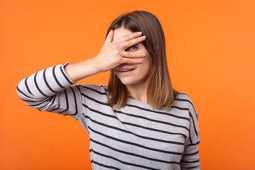 Portrait of curious cheerful woman with brown hair in long sleeve striped shirt standing, peeking through fingers at camera, spying looking for secret. indoor studio shot isolated on orange background