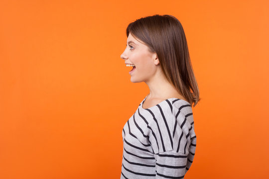 Side View Portrait Of Beautiful Amazed Woman With Brown Hair In Long Sleeve Striped Shirt Standing Surprised With Open Mouth, Looking To The Left. Indoor Studio Shot Isolated On Orange Background