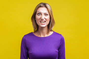 Fototapeta premium Portrait of beautiful emotional woman in tight purple dress standing with clenched teeth and angry grimace on face, pretending to be aggressive. indoor studio shot isolated on yellow background
