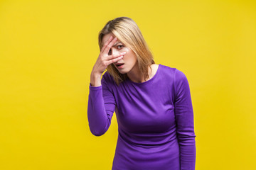 Portrait of curious young woman in elegant tight purple dress standing looking through fingers with...