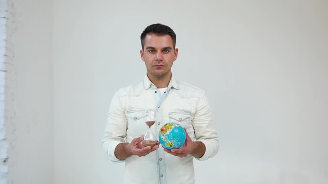 Handsome focused young well-dressed man standing on white background and posing on camera with small globe and sand clock in his hands