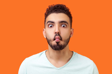 Portrait of funny silly brunette man with beard in casual white t-shirt looking at camera and making fish face with lips and big eyes, fooling around. indoor studio shot isolated on orange background - obrazy, fototapety, plakaty