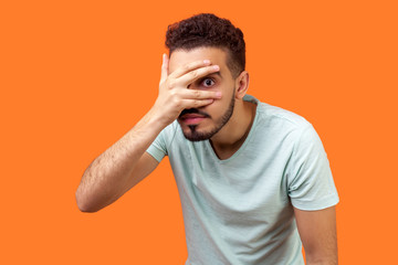 Portrait of curious brunette man with beard in white t-shirt covering face, looking through fingers...