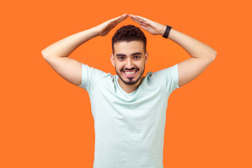Fototapeta na wymiar I'm safe. Portrait of cheerful brunette man with beard and toothy smile in casual white t-shirt standing with hands over head, making roof gesture. indoor studio shot isolated on orange background