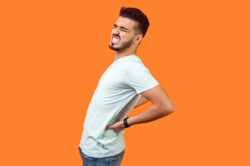 Fototapeta na wymiar Spine problems. Portrait of brunette man in casual white t-shirt holding his back, having sudden lower back pain, pinched nerve or kidney inflammation. indoor studio shot isolated on orange background