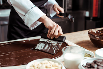 cropped view of chocolatier holding cake scraper near melted dark chocolate and milk