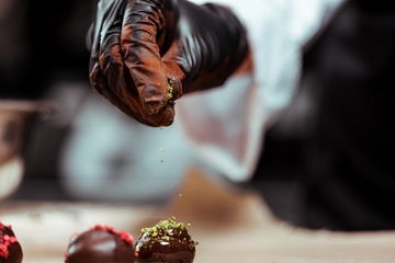cropped view of chocolatier in black latex glove adding green pistachio powder on fresh made candies