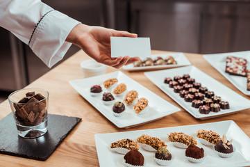 cropped view of chocolatier holding blank card in hand near tasty chocolate candies on plates