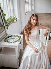 A beautiful bride in a white dress is sitting on a chair near the window and holding a wedding...