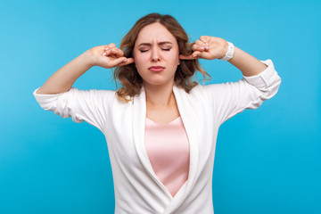 Fototapeta premium Don't want to listen! Portrait of resentful girl with wavy hair in white jacket standing covering ears and closed eyes in displeasure, frustrated about what she hear, ignoring loud noise. studio shot