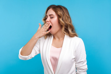 Portrait of fatigued woman with wavy hair in white jacket yawning feeling sleepy and tired, covering mouth with hand, exhausted of overwork, sleep disorder. studio shot isolated on blue background