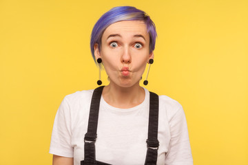 Portrait of amusing hipster girl with violet short hair in denim overalls making fish face with goofy comical expression, silly grimace, having fun. indoor isolated on yellow background, studio shot