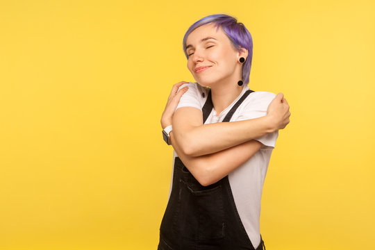 Portrait of beautiful hipster girl with violet short hair in denim overalls embracing herself and smiling from happiness and pleasure, self-love concept. isolated on yellow background, studio shot