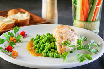 White dish with a portion of fried chicken breast and broiled green peas, cut white bread and a glass with thin slices of cucumbers and carrot. Nice festive service on the dark background.