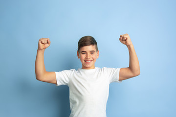 Celebrating win, happy. Caucasian boy portrait isolated on blue studio background. Beautiful teen male model in white shirt posing. Concept of human emotions, facial expression, sales, ad.