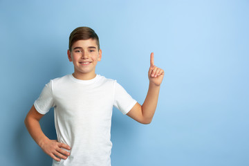 Naklejka premium Smiling and pointing up. Caucasian boy portrait isolated on blue studio background. Beautiful teen male model in white shirt posing. Concept of human emotions, facial expression, sales, ad.