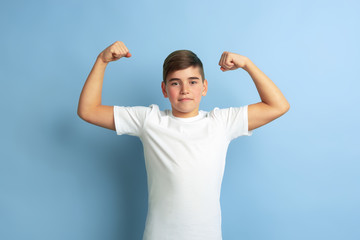 Celebrating win, happy, strong. Caucasian boy portrait isolated on blue studio background. Beautiful teen male model in white shirt posing. Concept of human emotions, facial expression, sales, ad.