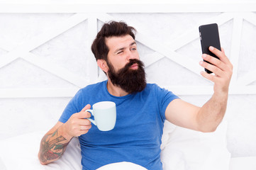 Selfie kiss. Bearded man take selfie in bed. Hipster smile to selfie camera in mobile phone. Enjoying selfie session. Morning coffee routine. Modern life. New technology. Social network