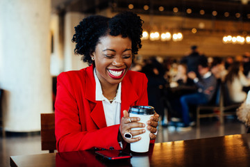 Portrait of a laughing young woman sitting in a cafe holding paper coffee cup with plastic lid