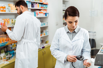 Pharmacist scanning jar of pills with barcode scanner with colleague at background