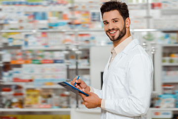 Selective focus of smiling pharmacist with clipboard looking at camera in apothecary