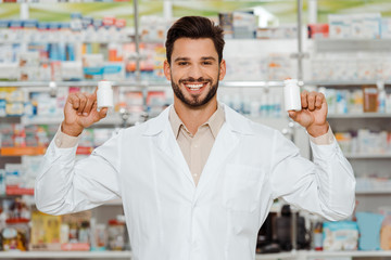 Handsome pharmacist smiling at camera and holding jars with pills