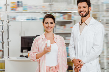 Fototapeta na wymiar Smiling woman with jar of pills beside pharmacist with drugstore showcase at background