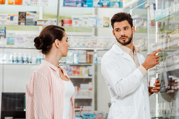 Fototapeta na wymiar Druggist with pills pointing with finger at shelves with medicaments to customer