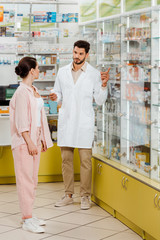 Fototapeta na wymiar Pharmacist holding medicament and pointing at pharmacy showcase to customer