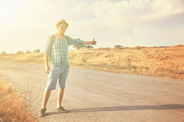 Hitchhiker with backpack in summer hat, light shirt, shorts on the road in tropical country in Sunny weather at autumn
