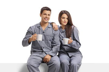 Young couple in pyjamas holding coffee mugs and sitting on a panel
