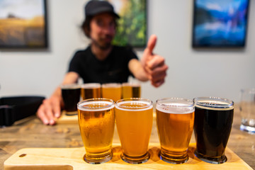 selective focus Flight of Craft of four different Beers glasses on Wooden Tray, happy caucasian man giving thumb sign finger gesture in the background