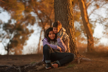 A big sister and young brother sitting near a tree on a hill and hugging each other while enjoying...