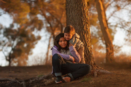 A big sister and young brother sitting near a tree on a hill and hugging each other while enjoying a sunset.