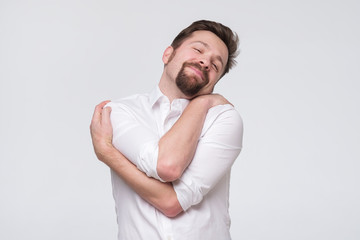 PLeased handsome bearded young man hugging himself with closed eyes and smile enjoying.